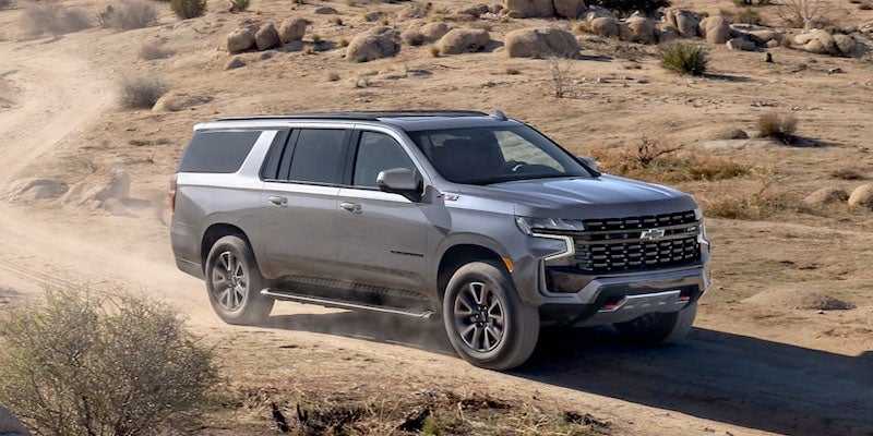 A 2022 Chevrolet Suburban driving along a rugged dirt road surrounded by trees and natural scenery.